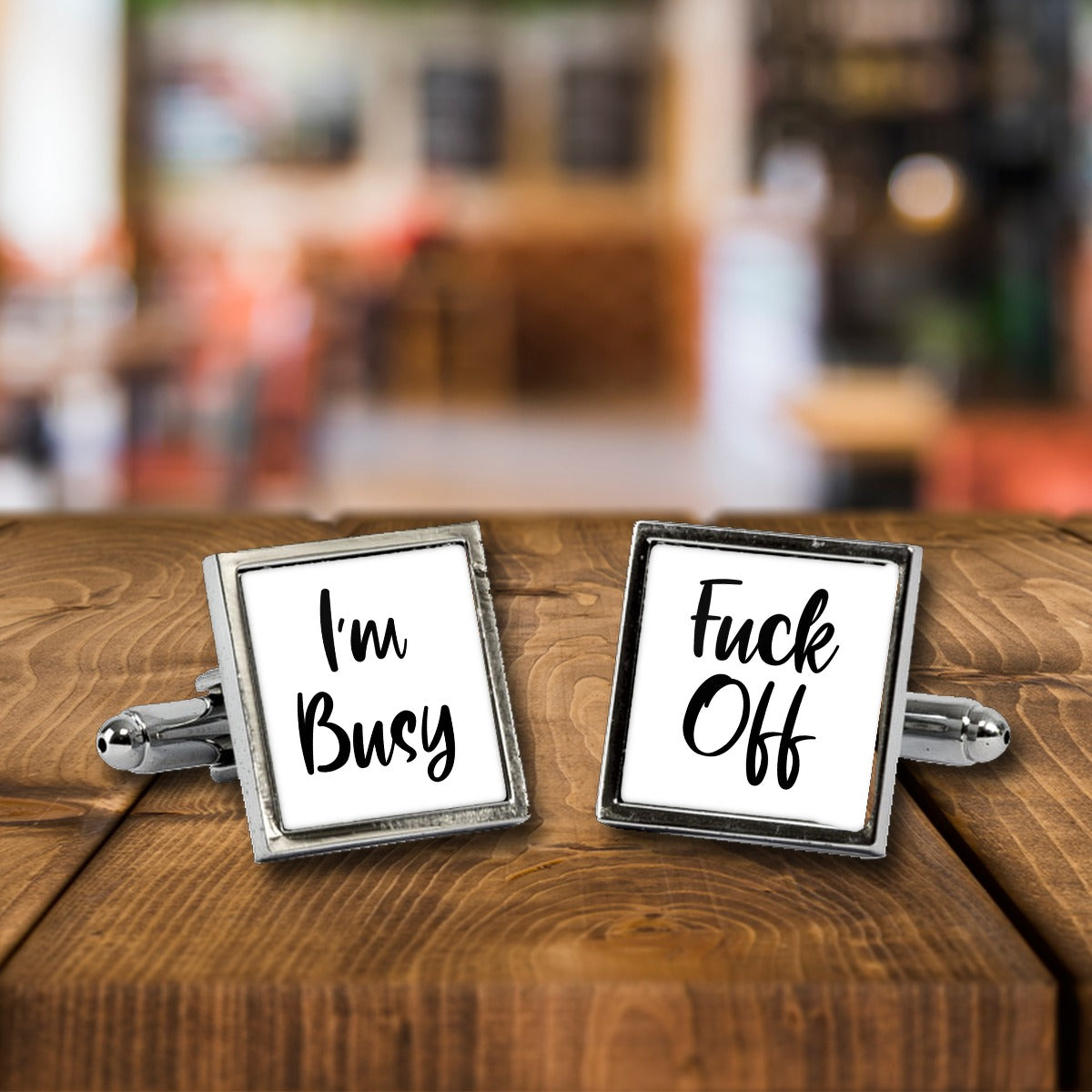 I'm Busy Fuck Off Cufflinks – Rude Novelty Cufflinks for Men – White or Black Print, Chrome or Rose Gold – I'm Busy Fuck Off cufflinks, a chrome square pair with white print on a wooden bar table