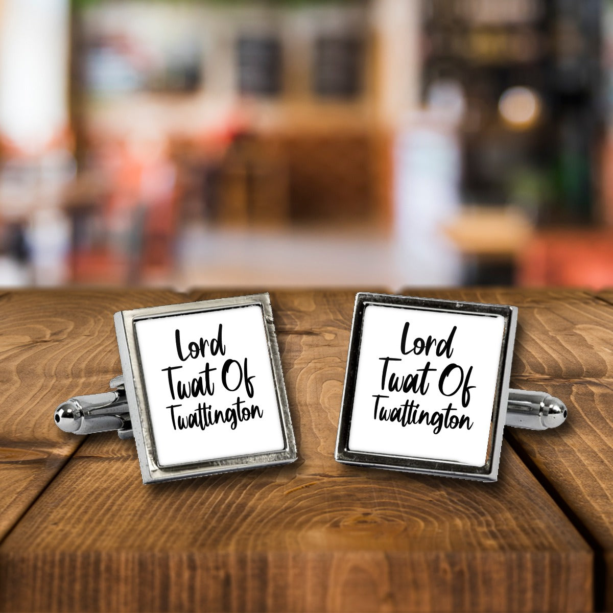 Lord Twat of Twattington Cufflinks – Rude Novelty Cufflinks for Men – White or Black Print, Chrome or Rose Gold – Lord Twat of Twattington cufflinks, a chrome square pair with white print on a wooden bar table