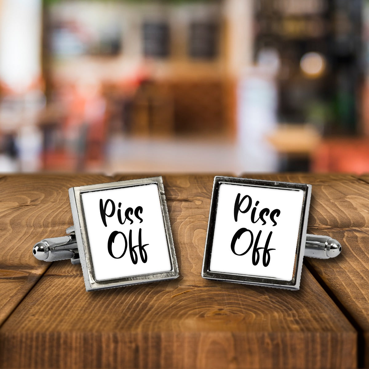 Piss Off Cufflinks – Rude Novelty Cufflinks for Men – White or Black Print, Chrome or Rose Gold – Piss Off cufflinks, a chrome square pair with white print on a wooden bar table