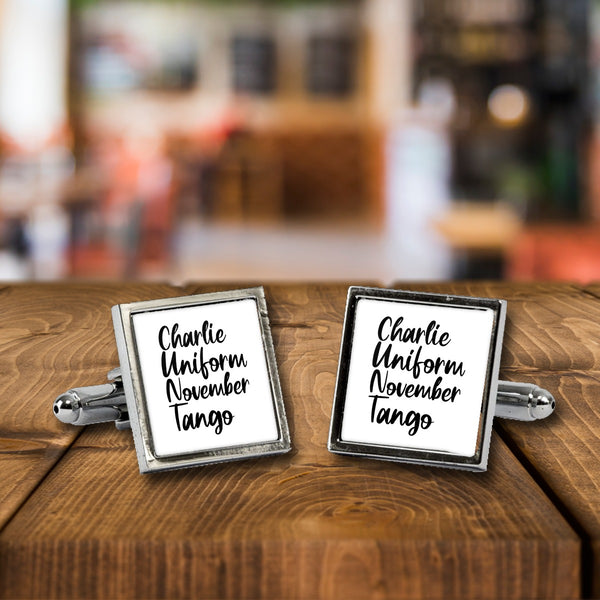 Charlie Uniform November Tango cufflinks, a chrome square pair with white print on a wooden bar table