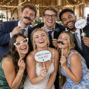 Wedding guests laughing with personalised photo booth props at a barn reception
