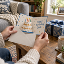 Personalised pop-up 3D birthday card sliding out of its envelope beside a wrapped present