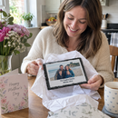 Personalised photo slate, being unwrapped from tissue paper by a smiling woman at a kitchen table on Mother's Day, with flowers nearby