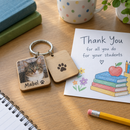 Personalised double-sided keyring: a personalised square double-sided wooden keyring printed with a photo of a cat and the name 'Mabel' next to a thank-you card for a teacher on a classroom desk