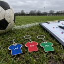 Personalised double-sided keyring: a row of personalised T-shirt shaped wooden keyrings in different team colours with names 'Leo', 'Ava', 'Max' laid out on a grassy pitch sideline next to a football