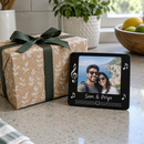 Personalised musical photo magnet: a gift-wrapped present beside a personalised black musical photo magnet showing a couple's holiday photo with the names 'Sam & Priya' on a kitchen worktop