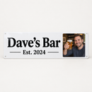 Personalised metal sign printed with bold text Dave's Bar and Est. 2024 alongside a small photo of a smiling man raising a pint glass with no logos, on a plain background