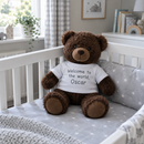 Personalised teddy bear: a dark brown plush teddy bear wearing a white t-shirt printed with 'Welcome to the world, Oscar' sitting in a white cot in a calm UK nursery with grey bedding