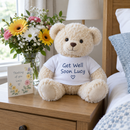 Personalised teddy bear: a cream teddy bear wearing a white t-shirt printed with the words 'Get Well Soon Lucy' sitting on a bedside table next to flowers and a card, gentle daylight