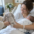 Personalised bunny rabbit soft toy: a grey plush bunny rabbit soft toy printed with the words 'Hello little Arthur' being handed to a new mum in a hospital-style room with a baby in her arms, gentle light