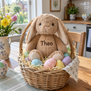 Personalised bunny rabbit soft toy: a light brown plush bunny rabbit printed with the name 'Theo' sitting in an Easter basket with pastel eggs on a kitchen table in a UK home, spring morning light