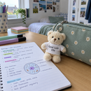Personalised mini teddy bear keyring: a cream mini teddy bear keyring wearing a white t-shirt printed with 'Good Luck Ella' clipped to a pencil case on a desk with revision notes in a UK teenager's bedroom