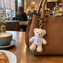Personalised mini teddy bear keyring: a woman's handbag on a café table with a cream mini teddy bear keyring clipped to the strap wearing a t-shirt printed 'Jess & Callum' and a small heart