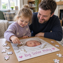 A toddler and her dad doing a personalised kids jigsaw on a low table showing a photo of a new baby and the text 'Big Sister Molly', cosy British living room, daylight