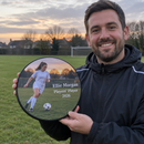 Personalised award plaque, held by a smiling coach in a tracksuit on a grass pitch at dusk