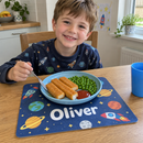 Personalised kids placemat, under a plastic plate of fish fingers and peas on a kitchen table with a smiling five-year-old boy