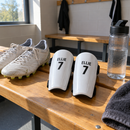 Pair of personalised white shin pads with boots and a water bottle on a changing room bench