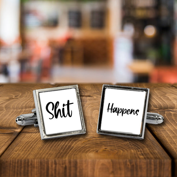 Shit Happens cufflinks, a chrome square pair with white print on a wooden bar table